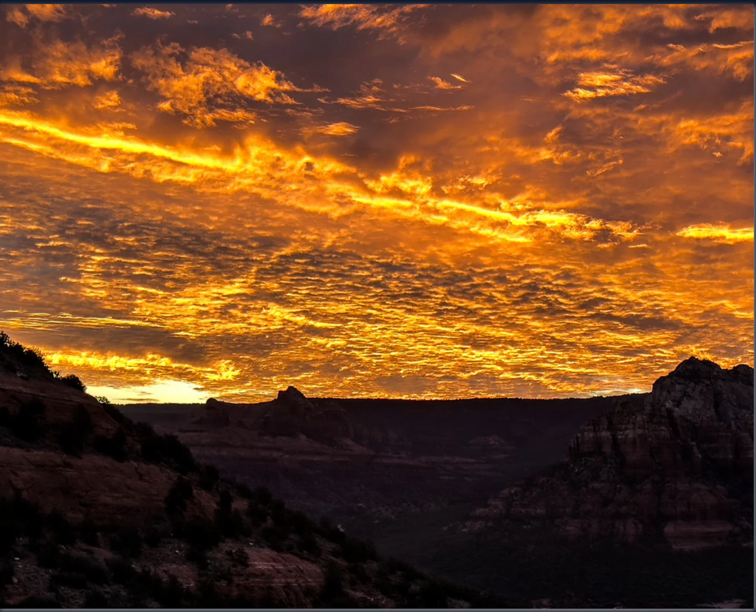 Dramatic golden-orange sunrise illuminating textured clouds above the dark red rock landscape of Sedona, Arizona.