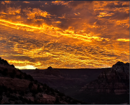 Dramatic golden-orange sunrise illuminating textured clouds above the dark red rock landscape of Sedona, Arizona.
